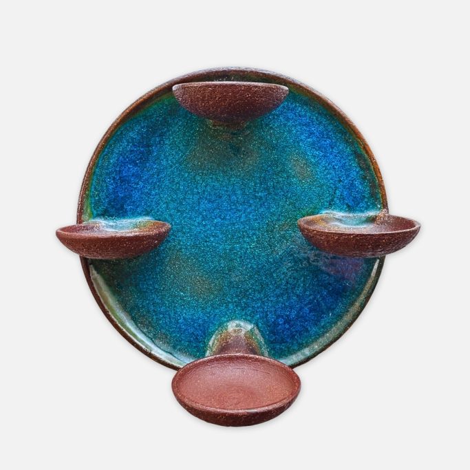 Ein runder Wandkerzenhalter aus Keramik mit vier Halterungen für Stumpenkerzen. Das Stück ist blau glasiert, die Halter unglasiert in braun.
